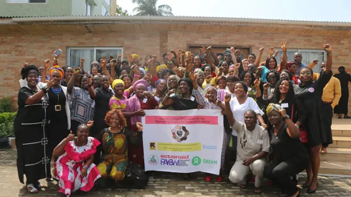 A large group of women raising their hands in celebration. In front of them is a banner reading "The Official National Launch of The Feminist Humanitarian Network in Liberia."