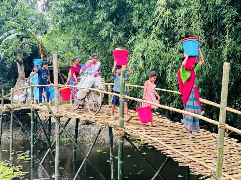 a group of women and children carry dry ration and hygiene kits across a bridge in India.
