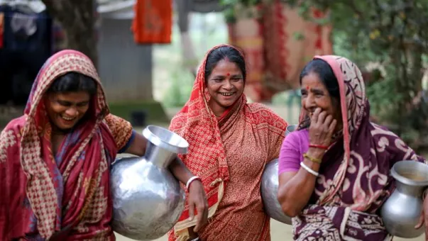 Three women smile and talk as they walk holding silver water pots.